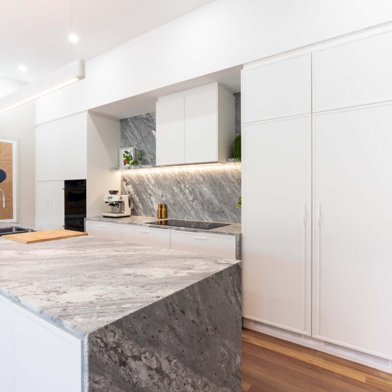Modern kitchen with large island benchtop. White thin shaker profiled cabinets with a bold grey marble benchtop and splashback. Large concealed double door fridge with matte white handles.