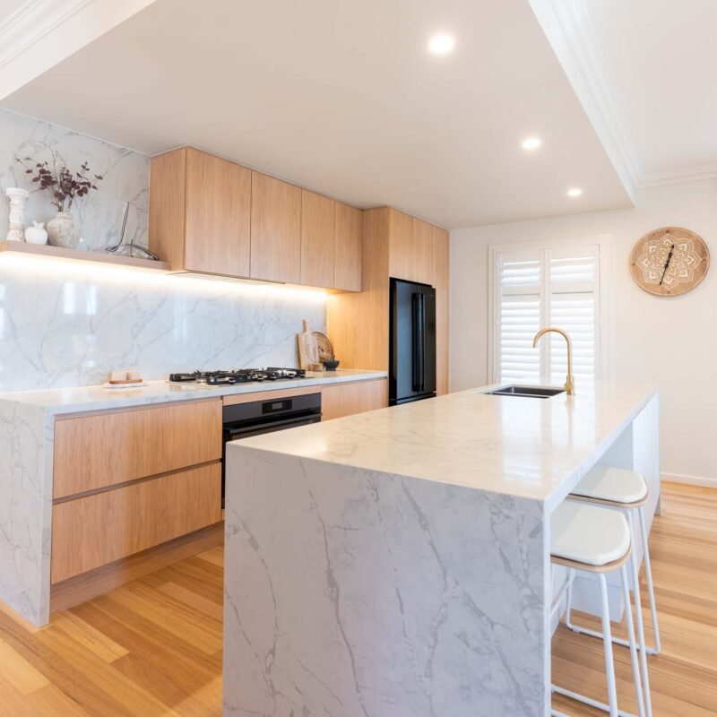 A modern classic kitchen with a large island benchtop. Featuring oak timber laminate finger pull cabinets and light marble benchtops with waterfall ends.
