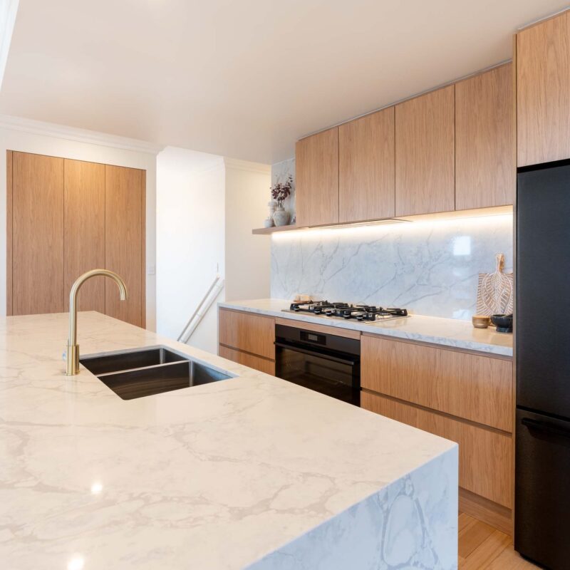 A modern classic kitchen with a large island benchtop. Featuring oak timber laminate finger pull cabinets and light marble benchtops with waterfall ends.