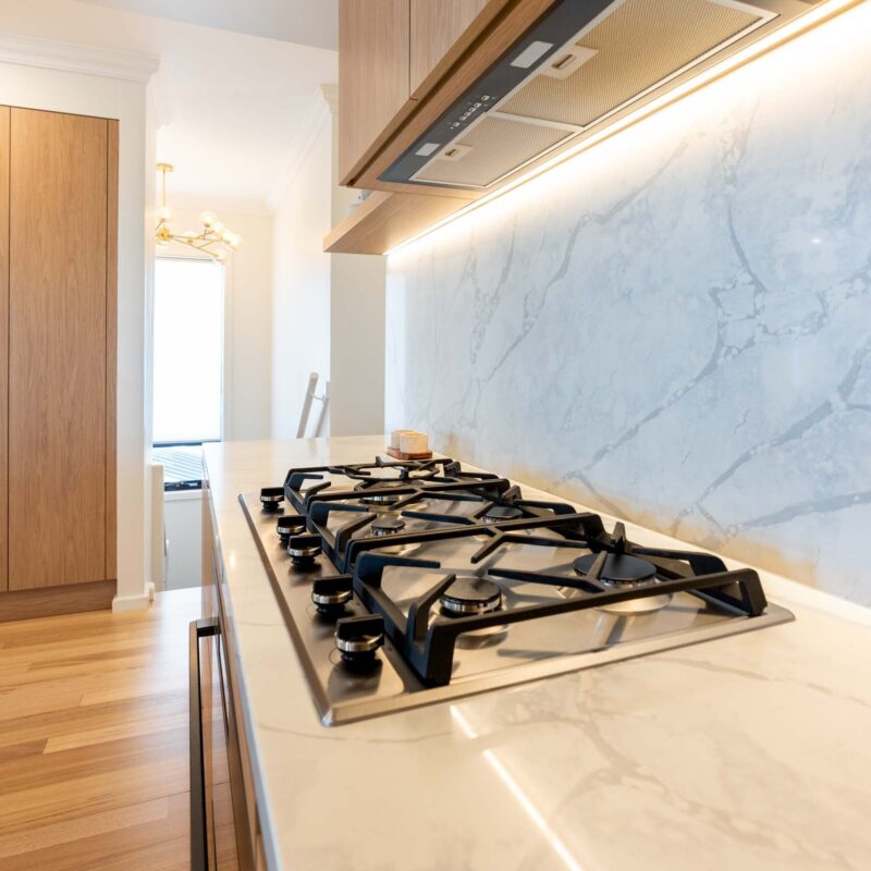 Kitchen with light marble benchtops and matching splashback. Featuring a 900 gas cooktop and timber laminate overhead cabinets with undermount LED strip lighting.