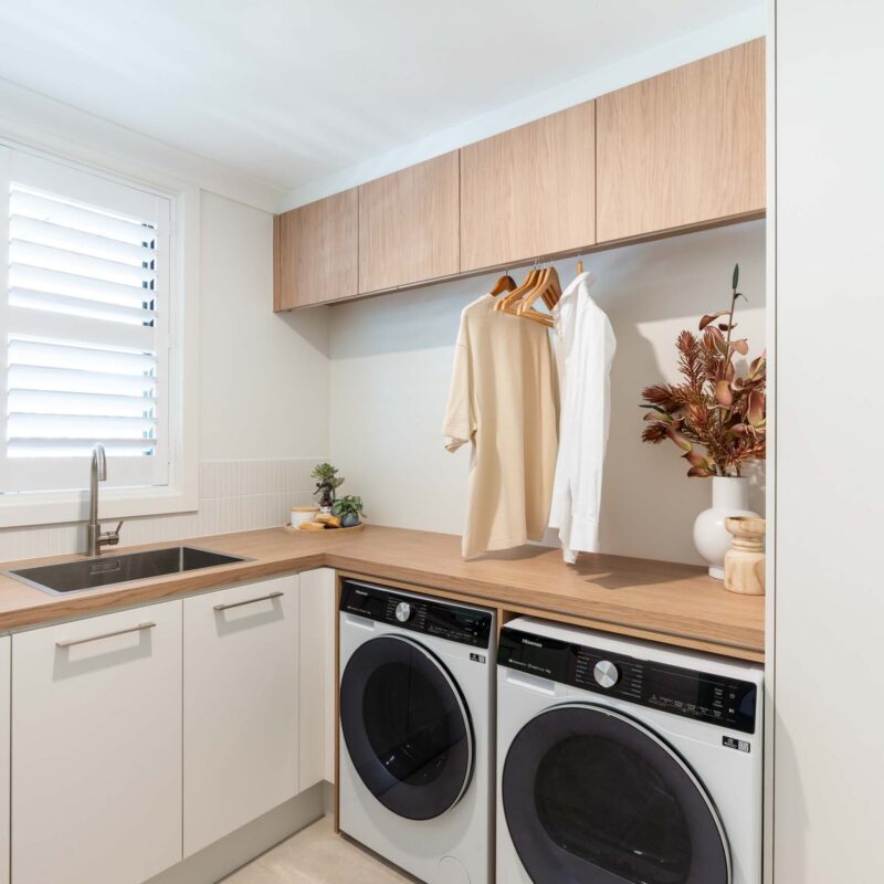 Laundry with a timber look laminate benchtops and white matte cabinets. Featuring a hanging rail and timber laminate over head cabinets. Under bench washing machine and dryer with a large broom cabinet to the right.