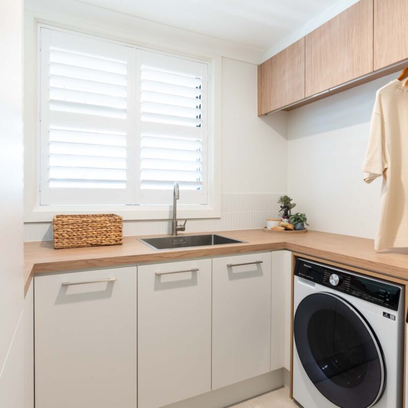 Laundry with a timber look laminate benchtops and white matte cabinets. Featuring a hanging rail and timber laminate over head cabinets. Under bench washing machine.