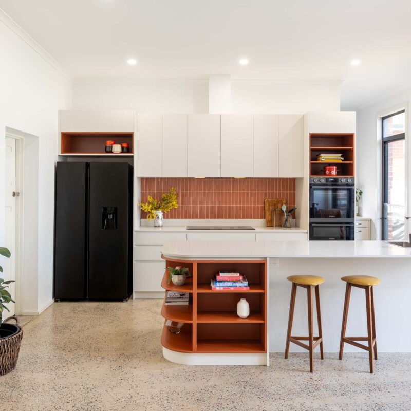 A mid-century modern kitchen featuring a mixture of matte white finger pull cabinets and burnt orange laminate open shelf cabinets. Includes a thin white engineered stone benchtop and a fluted orange tiled splashback.