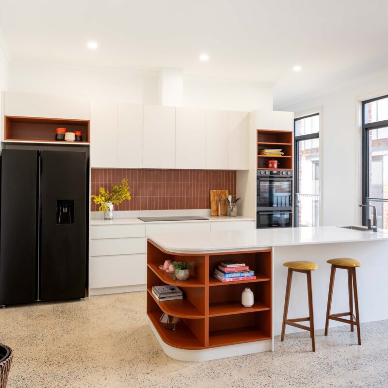 A mid-century modern kitchen featuring a mixture of matte white finger pull cabinets and burnt orange laminate open shelf cabinets. Includes a thin white engineered stone benchtop.