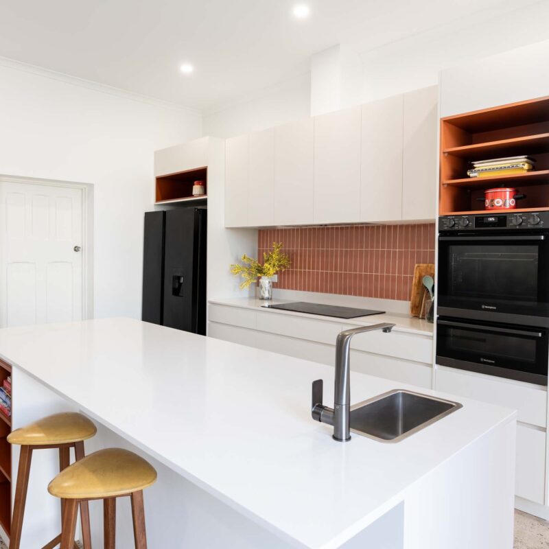 A mid-century modern kitchen featuring a mixture of matte white finger pull cabinets and burnt orange laminate open shelf cabinets. Includes a thin white engineered stone benchtop.