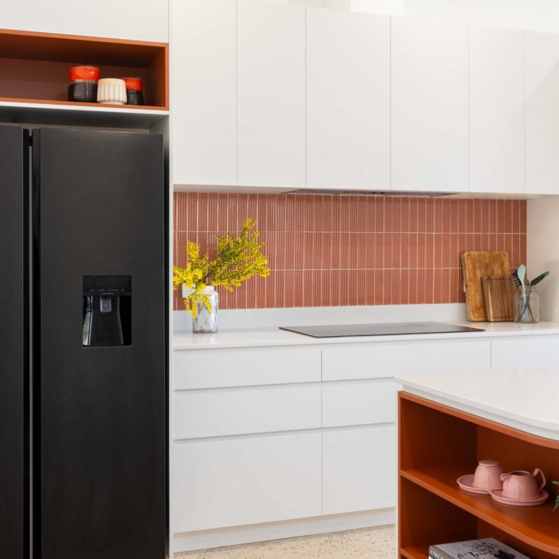 A mid-century modern kitchen featuring a mixture of matte white finger pull cabinets and burnt orange laminate open shelf cabinets. Includes a thin white engineered stone benchtop and a fluted orange tiled splashback.