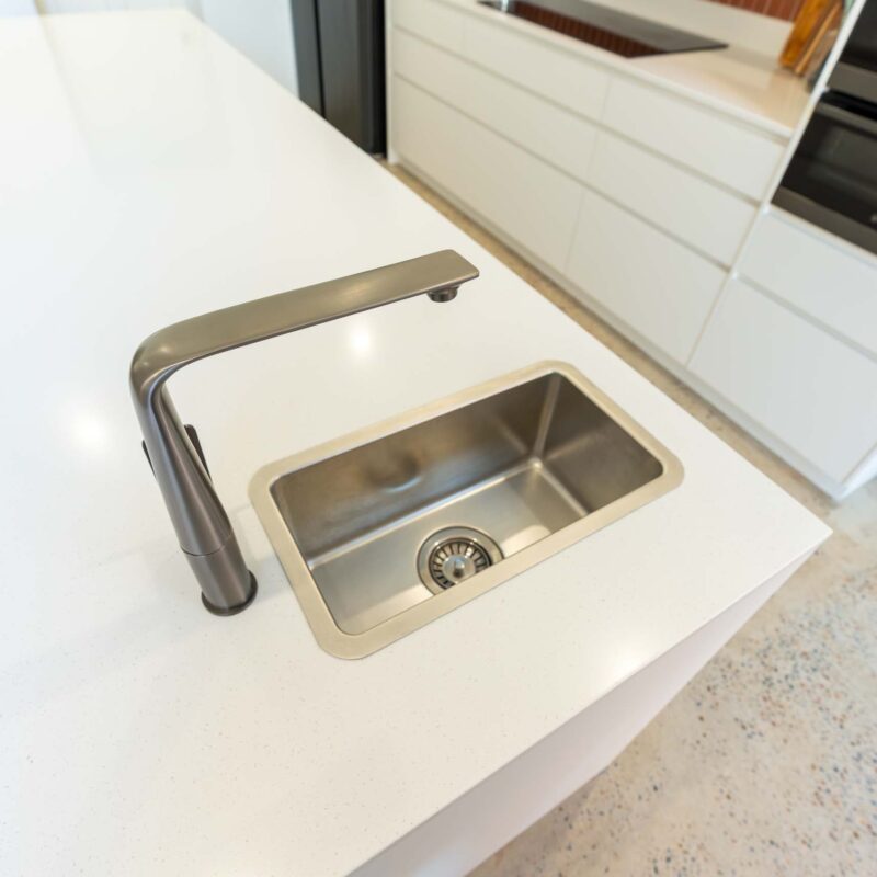 A small top mount stainless steel sink with gun metal grey mixer tap. Installed in a white stone benchtop.
