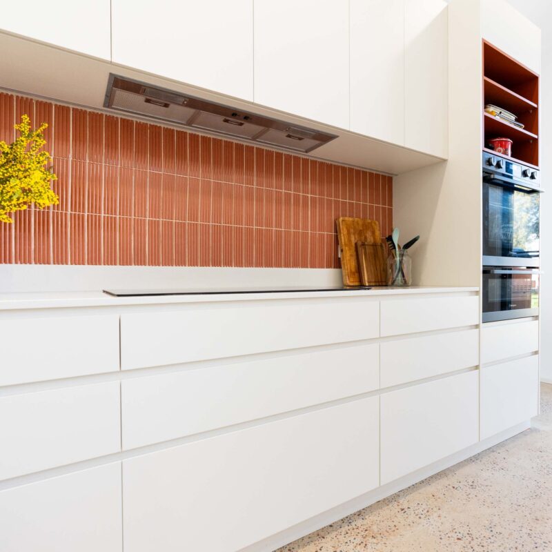 A mid-century modern kitchen featuring a mixture of matte white finger pull cabinets and burnt orange laminate open shelf cabinets. Includes a thin white engineered stone benchtop. Fluted orange feature tiled splashback.
