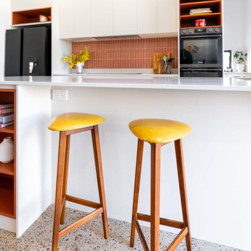 A mid-century modern kitchen featuring a mixture of matte white finger pull cabinets and burnt orange laminate open shelf cabinets. Includes a thin white engineered stone benchtop.