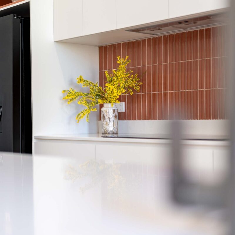 Kitchen featuring matte white finger pull cabinets. Includes a thin white engineered stone benchtop and a fluted orange feature tile splashback.