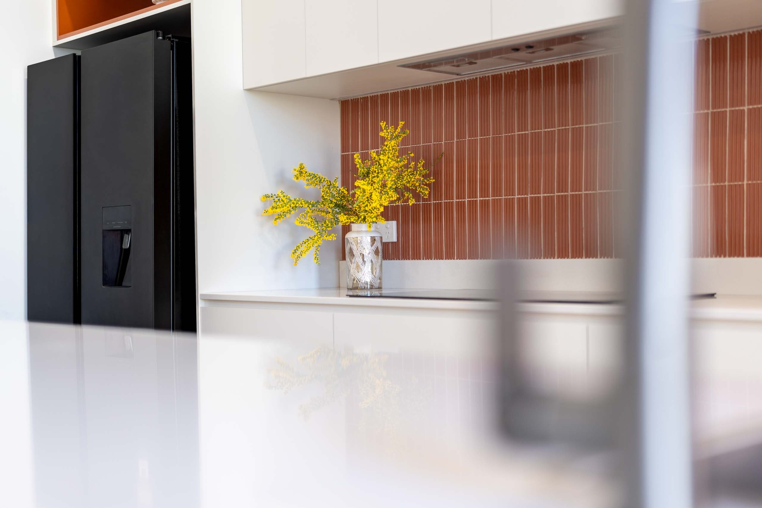 Kitchen featuring matte white finger pull cabinets. Includes a thin white engineered stone benchtop and a fluted orange feature tile splashback.