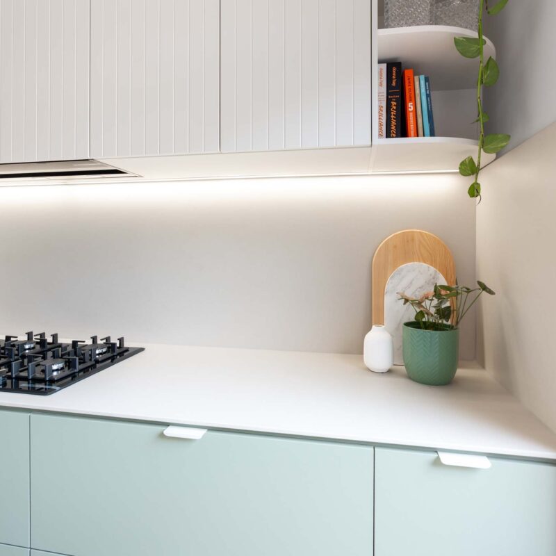 Two tone kitchen with green base cabinets and slatted overhead cabinets. Features a curved open shelf overhead cabinet. Includes a gas cooktop and integrated rangehood.