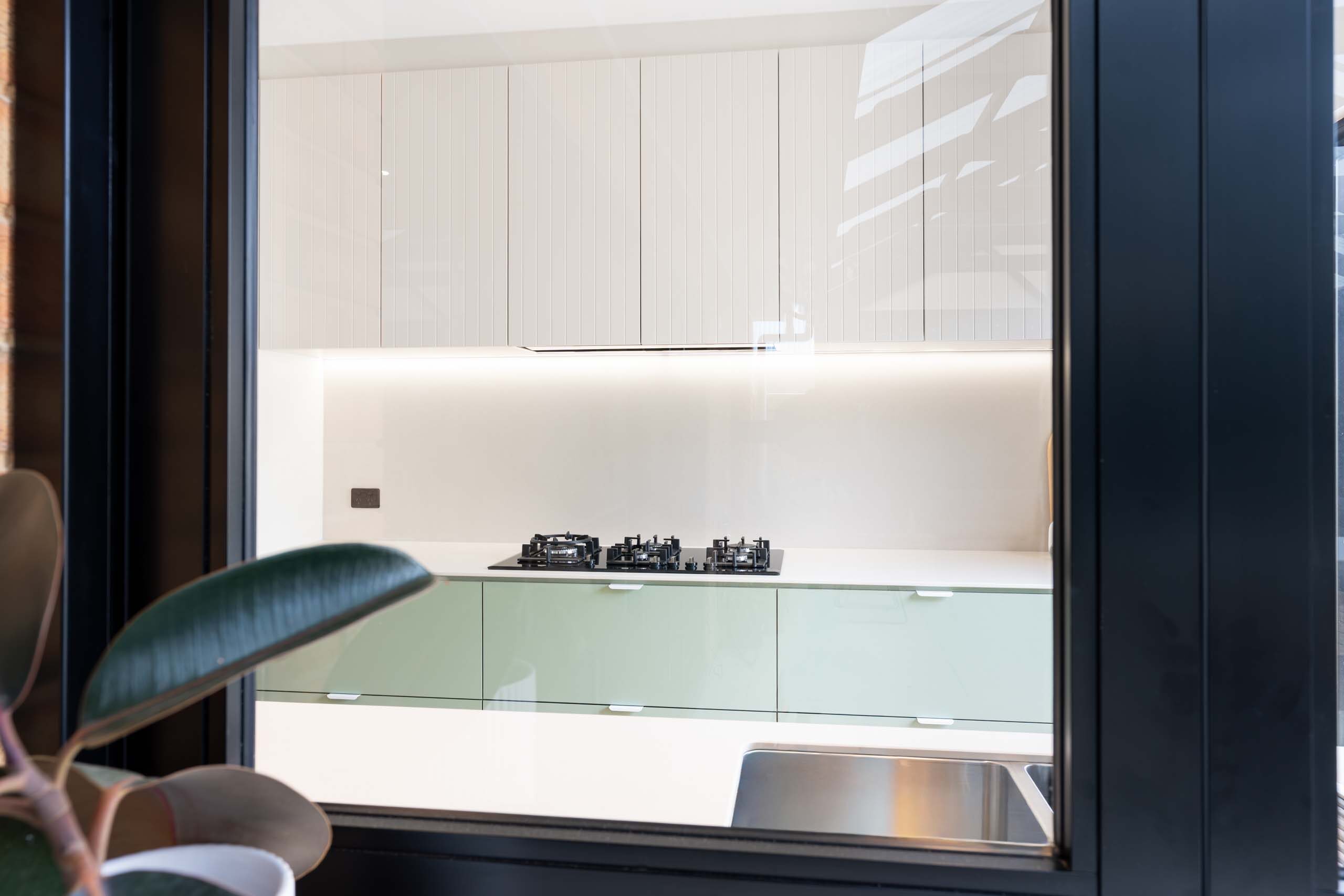 Looking through the kitchen window into a two tone kitchen featuring green base cabinet and slatted white overhead cabinets.