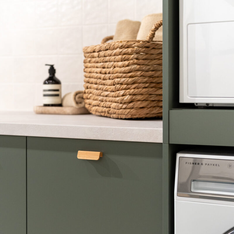 Matte green laundry showcasing a washing machine and dryer stack. Light laminate benchtop with a light square tiled splashback.