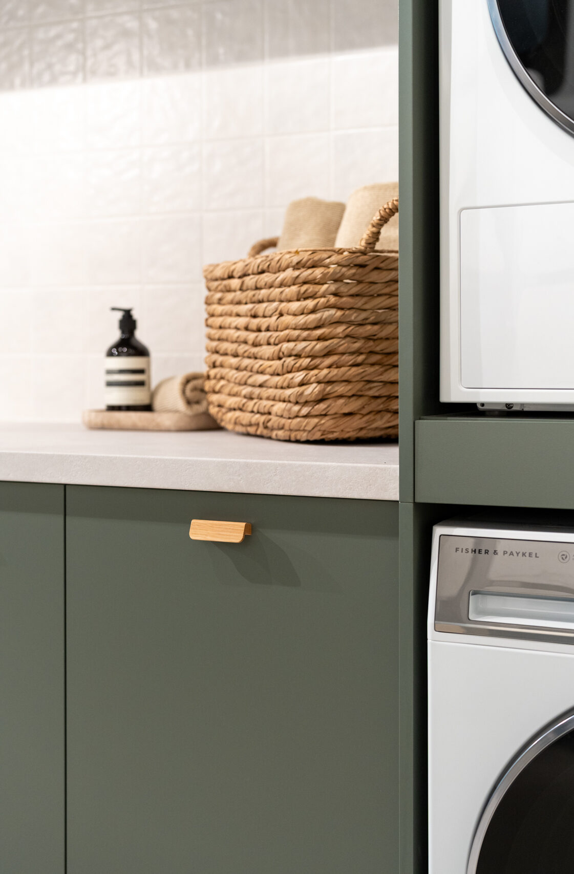 Matte green laundry showcasing a washing machine and dryer stack. Light laminate benchtop with a light square tiled splashback.