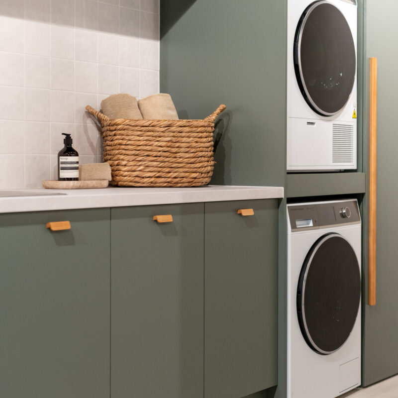 Matte green laundry showcasing a washing machine and dryer stack. Light laminate benchtop with a light square tiled splashback.