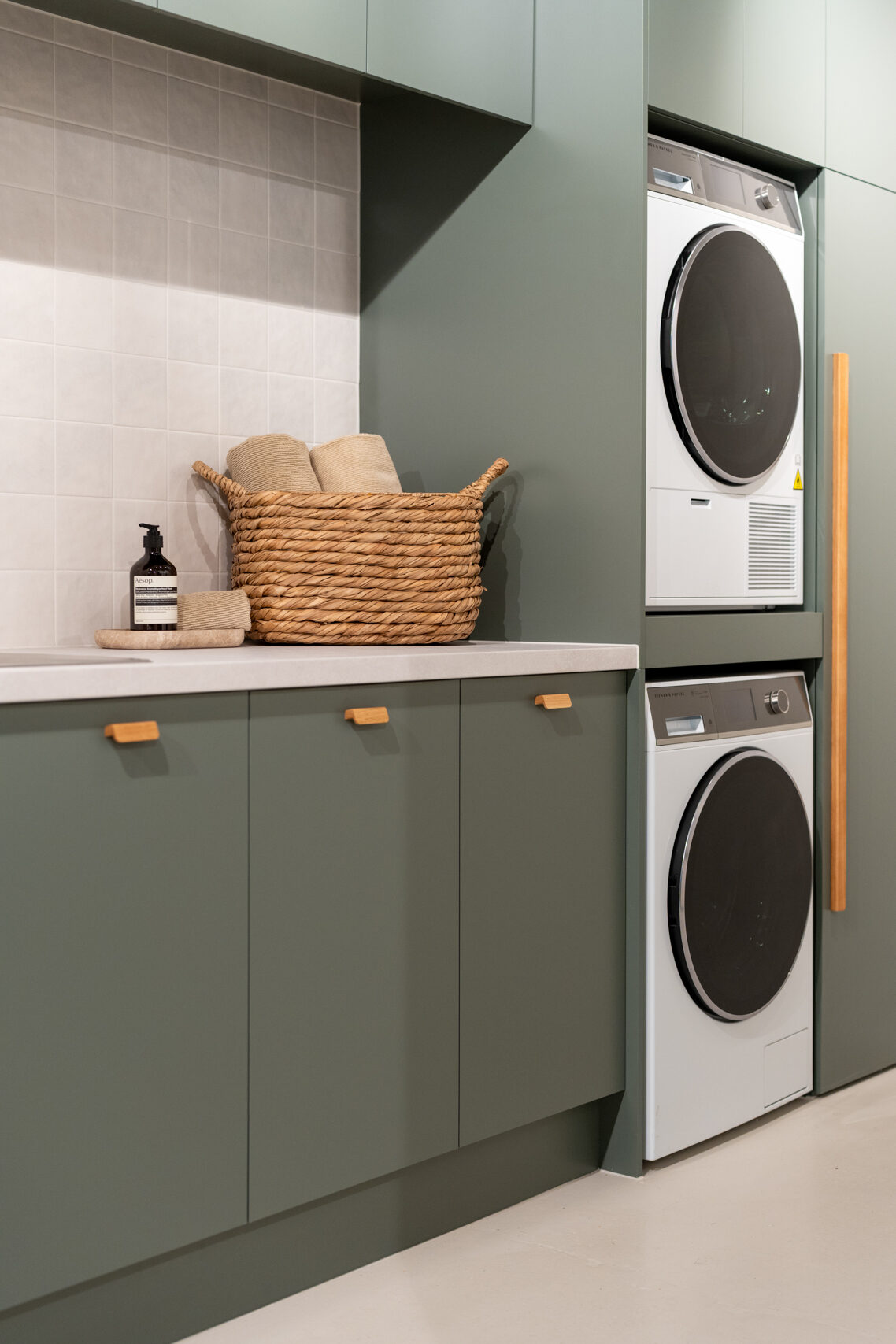 Matte green laundry showcasing a washing machine and dryer stack. Light laminate benchtop with a light square tiled splashback.