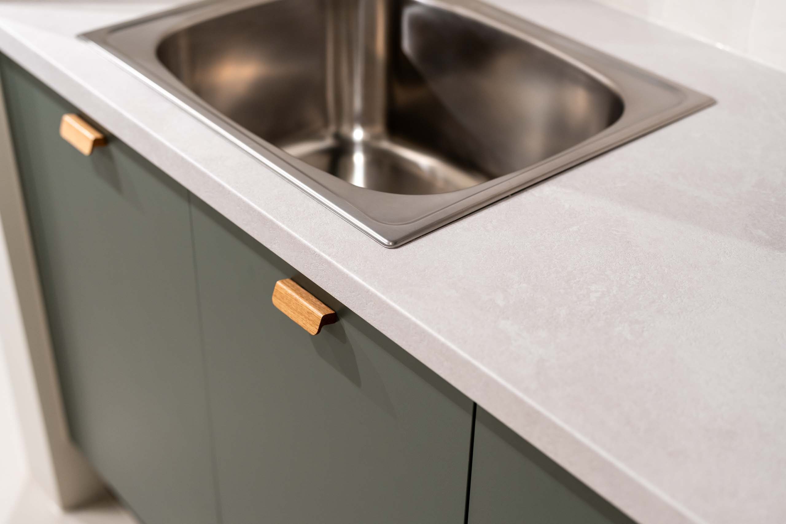 Laundry display featuring matte green cabinets with small timber handles. A light laminate benchtop.