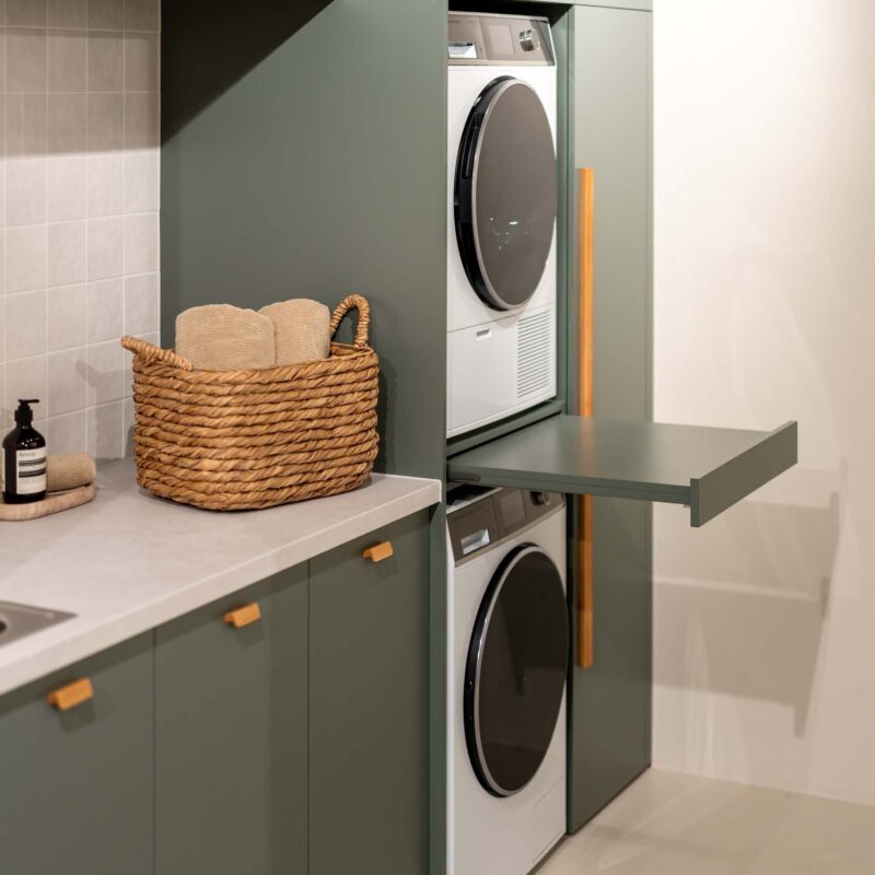 Matte green laundry showcasing a washing machine and dryer stack. Light laminate benchtop with a light square tiled splashback.