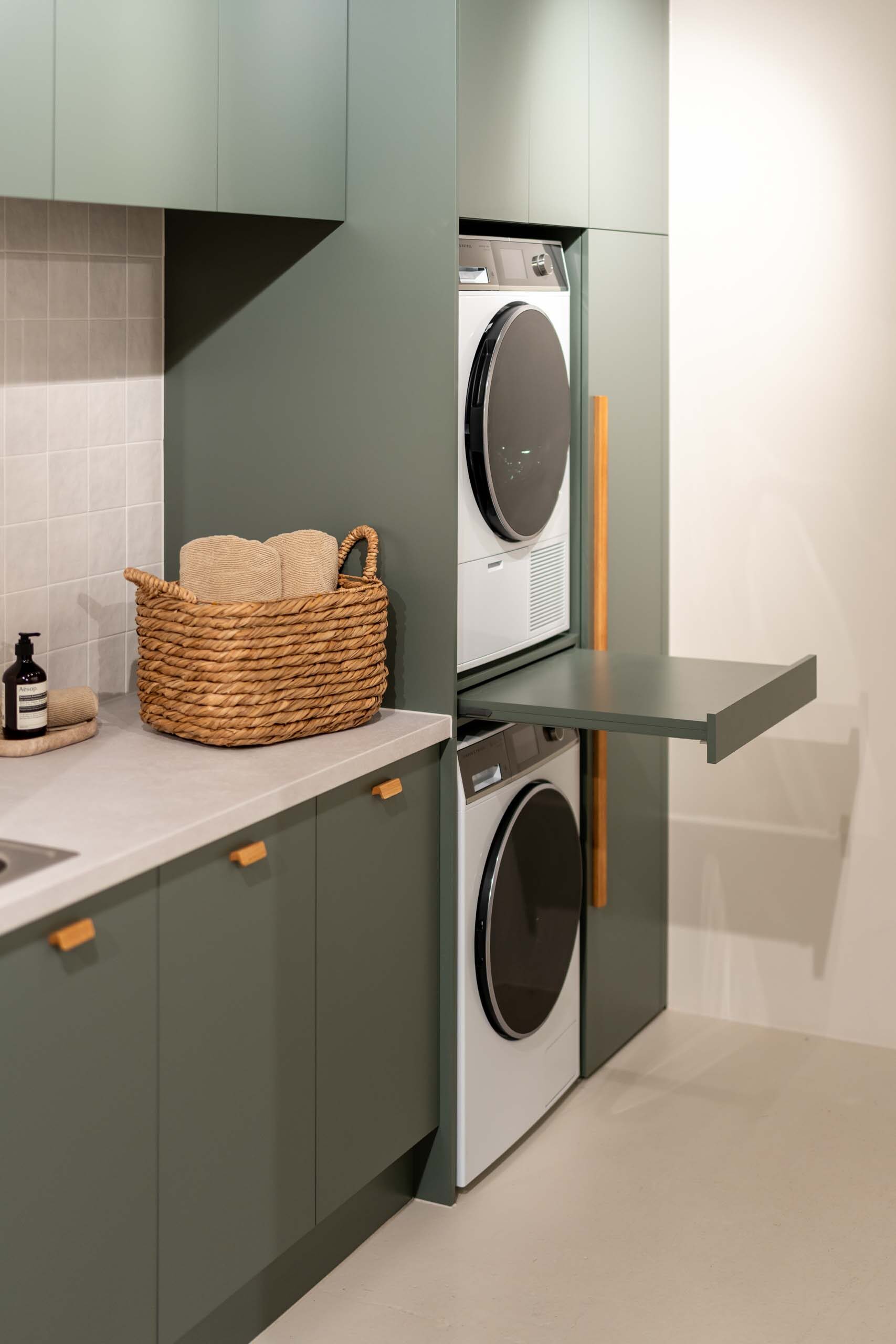 Matte green laundry showcasing a washing machine and dryer stack. Light laminate benchtop with a light square tiled splashback.